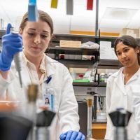 Two researchers in lab coats working in a laboratory, one using a pipette while the other observes.